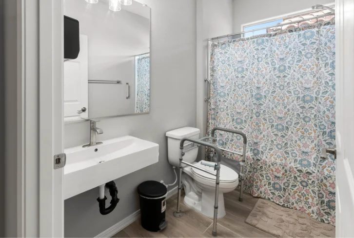Bathroom with toilet safety rails, shower and wall-mounted sink at Sunrise Haven Senior Homes